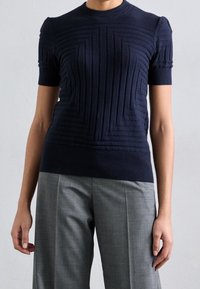 Jersey de punto acanalado en azul marino de manga corta con patrones geométricos texturizados y cuello redondo, combinado con pantalones ajustados en gris claro.