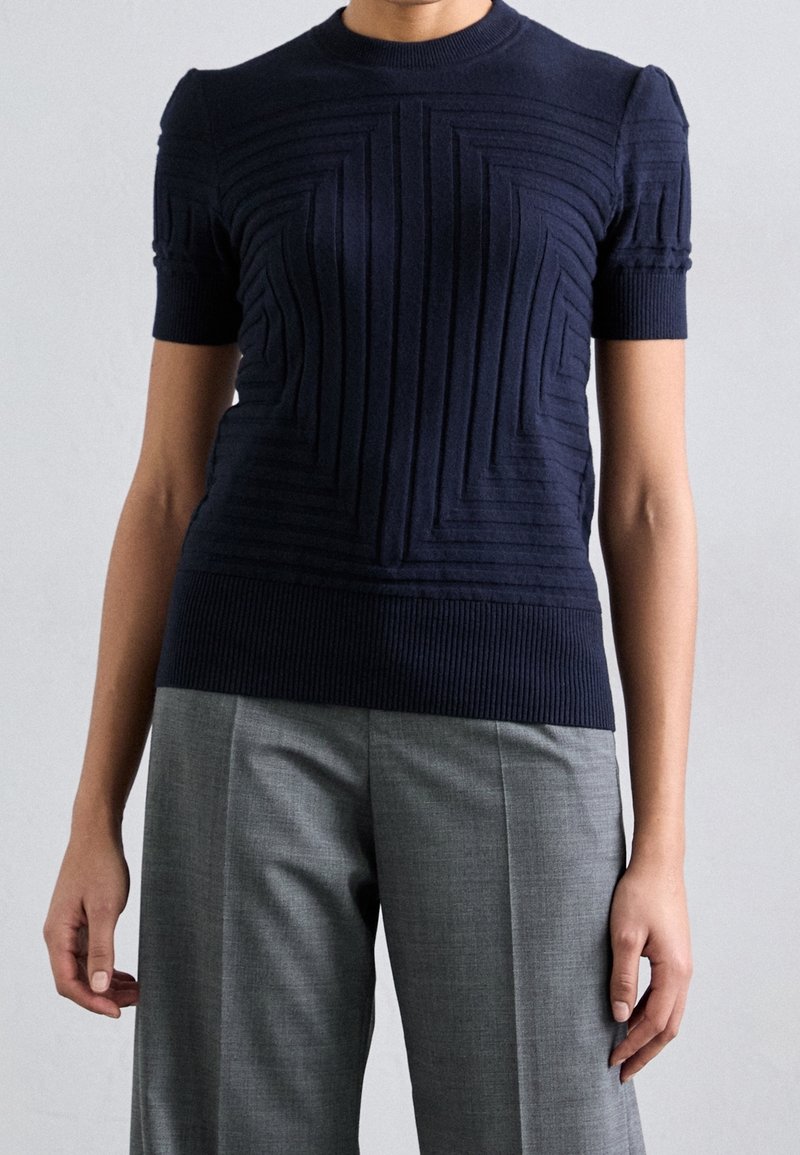 Jersey de punto acanalado en azul marino de manga corta con patrones geométricos texturizados y cuello redondo, combinado con pantalones ajustados en gris claro.