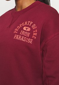 Felpa bordeaux con scollo a giro, caratterizzata dal testo "PROPRIETÀ DELL'IRON PARADISE" in arancione sbiadito. Tessuto morbido, polsini a coste.