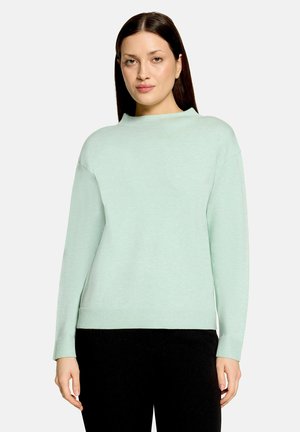 Femme aux cheveux lisses et foncés portant un pull vert clair à manches longues et un pantalon noir sur un fond clair uni.