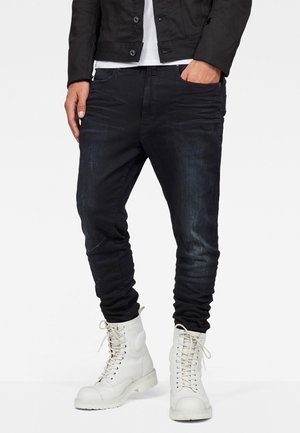 Mann trägt schwarze Slim-Fit-Jeans, weiße Schnür-Kampfboots, schwarze Jacke und weißes Hemd, steht mit einer Hand in der Tasche.
