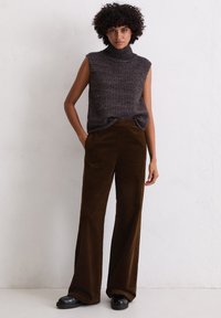 Mouwloze, hoge hals, donkergrijze gebreide trui gecombineerd met wijdvallende, bruine corduroy broeken. Zwarte plateau schoenen zichtbaar. Geweven stofdetails.