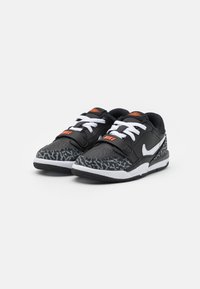 Black sneakers with white laces and strap, featuring a gray textured pattern on the toe. Small orange logo accents near the tongue.