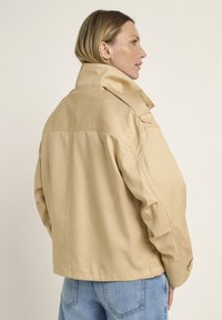 Beige cotton-blend jacket with a high collar, relaxed fit, and visible stitching. Features side pockets and a smooth texture.