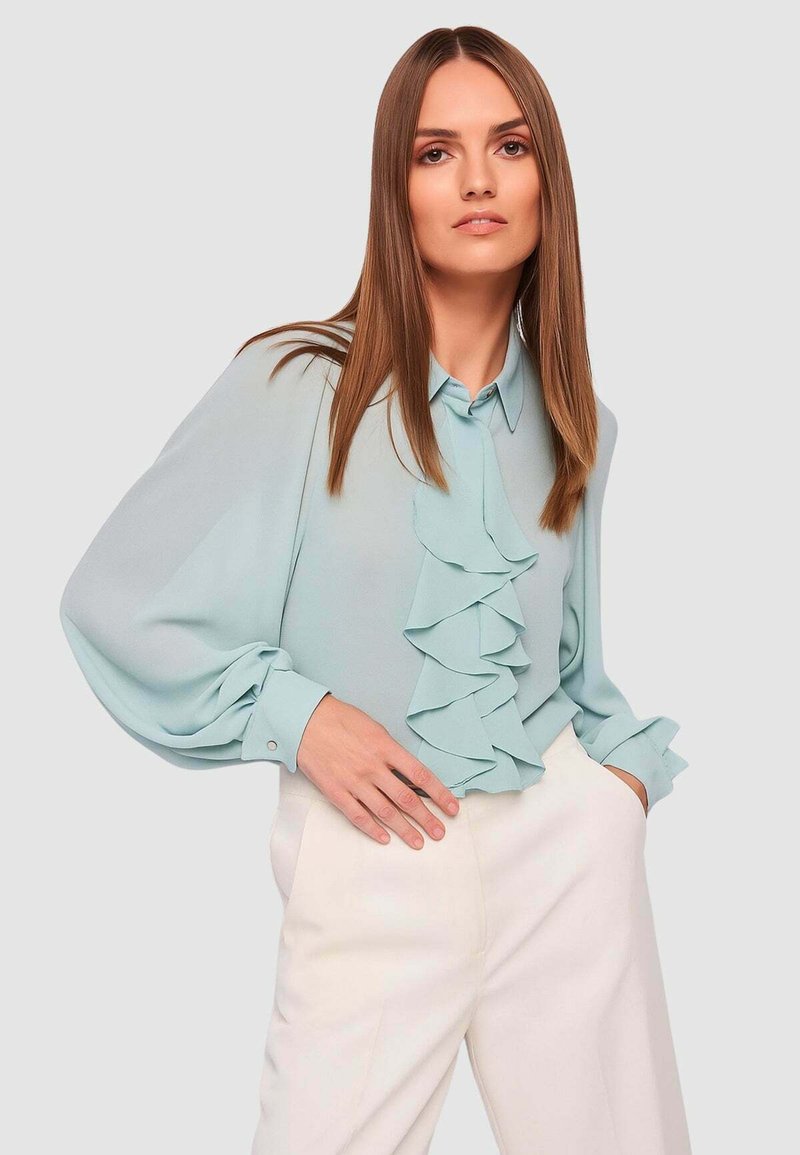 Camicia azzurro chiaro in tessuto morbido con una fila di balze sul davanti. Presenta un colletto con bottoni e maniche a sbuffo. Abbinata a pantaloni bianchi.