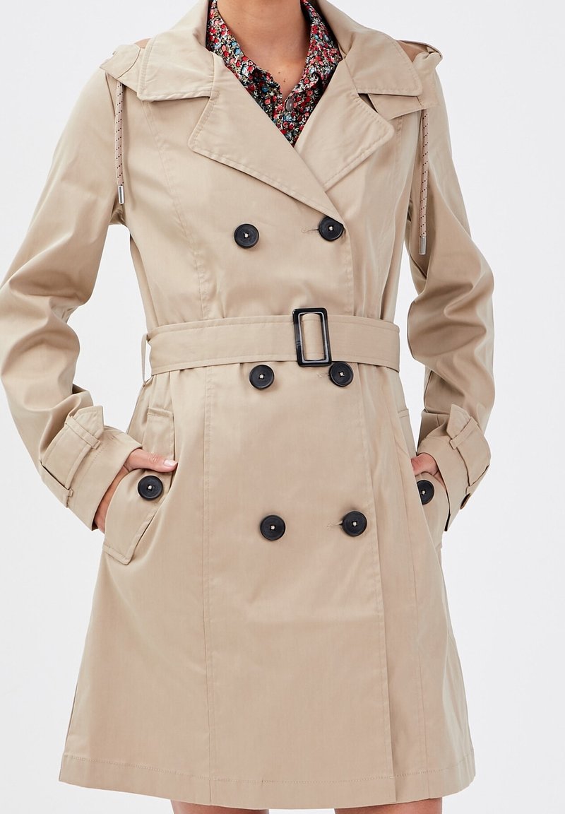 Beige double-breasted trench coat with black buttons and belt, worn over a red floral shirt, hands in pockets, on a person.