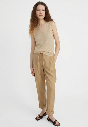 Junge Frau mit langen lockigen Haaren, die einen beigen ärmellosen Stricktop, braune Kordelhosen und schwarze Sandalen trägt, steht vor weißem Hintergrund.