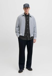 Chaqueta bomber gris con puños y dobladillo acanalados, camisa negra a cuadros, pantalones holgados negros y una gorra azul marino con marca blanca.