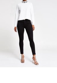 Frau, die ein weißes langärmliges Cropped-Top mit "AMOUR"-Kragen, schwarze enge Jeans und weiße Riemchen-High-Heels trägt, steht vor einem weißen Hintergrund.