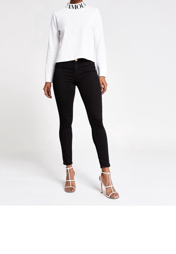 Frau, die ein weißes langärmliges Cropped-Top mit "AMOUR"-Kragen, schwarze enge Jeans und weiße Riemchen-High-Heels trägt, steht vor einem weißen Hintergrund.
