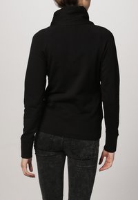 Svart sweatshirt med hög krage, långa ärmar och ribbad fåll. Texturen ser mjuk ut. Bärs med mörka, urtvättade jeans.