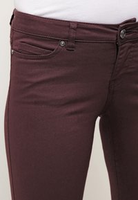 Jeans de denim burdeos con una textura suave, diseño de cinco bolsillos y herrajes en tono plateado. Presentan un estilo ajustado y un cierre con cremallera.