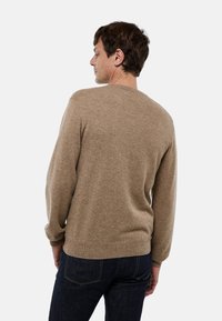 Homme faisant face à gauche, portant un pull à manches longues couleur marron clair et un jean bleu foncé devant un fond blanc uni.