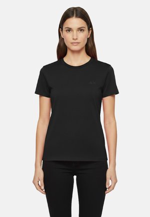 Donna con capelli castani lisci che indossa una maglietta aderente nera a maniche corte e pantaloni neri, in piedi davanti a uno sfondo semplice.