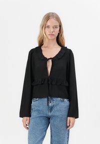 Black long-sleeve blouse with ruffled neckline and hem, featuring a tie closure at the front, paired with light blue jeans.