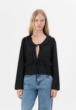Black long-sleeve blouse with ruffled neckline and hem, featuring a tie closure at the front, paired with light blue jeans.