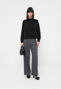 Una donna con capelli castani lisci indossa un blouse nero a maniche lunghe, pantaloni grigi a gamba larga, décolleté neri con fiocco e tiene una piccola borsa nera.