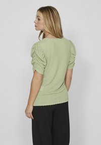 Lichtgroene textuur top met pofmouwen, voorzien van een ronde halslijn en een aansluitende pasvorm. Draagt met zwarte broeken.