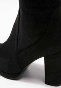 Černý semišový kotník na podpatku s robustním podpatkem, hladkým povrchem a ostrými výřezy na bocích pro zvýšení detailu a designového zájmu.