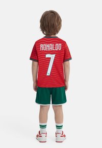 Παιδί με κόκκινη και πράσινη στολή ποδοσφαίρου, όρθιο με την πλάτη να φαίνεται, με τη φανέλα που γράφει «Ρονάλντο 7», λευκά παπούτσια και ριγέ κάλτσες.