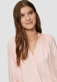 Lichtroze blouse met lange mouwen en een V-halslijn, met geplooide details bij de hals. Gemaakt van zachte stof, met een subtiele textuur.