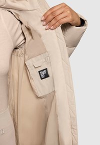 Beige quiltet jakke med en indre lomme, der har et logo. Glat tekstur, minimalistisk design og et varmt indrefor.