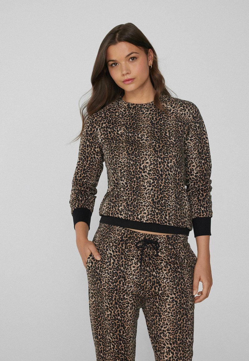 Mujer con cabello castaño largo vistiendo un conjunto de blusa y pantalones de leopardo a juego con puños y cinturilla negros, con las manos en los bolsillos.