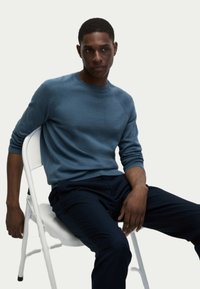 Blauer gestrickter Pullover mit gerippten Bündchen und Saum, getragen über dunklen Hosen, sitzend auf einem weißen Stuhl vor einem hellen Hintergrund.