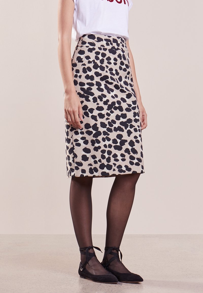 Femme portant une jupe beige à taches noires jusqu'aux genoux, des collants noirs transparents, des ballerines noires avec des rubans à la cheville, et un haut blanc imprimé.