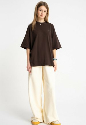 Młoda kobieta ubrana w luźny ciemnobrązowy T-shirt i szerokie kremowe spodnie, stoi na białym tle.