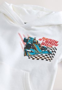 Balta džemperio su gobtuvu su priekine kišene. Ant jo yra mėlynas lenktyninio automobilio grafikas judesyje, su raudonomis detalėmis ir po juo raštuotas languotas vėliavos motyvas.