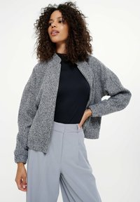 Kobieta z kręconymi włosami, ubrana w szaro-zapinany na zamek kurtkę, czarną bluzkę i jasnoszare, wysokie, szerokie spodnie, pozując z jedną ręką na biodrze.