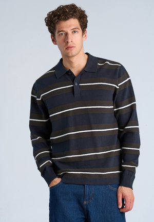 Young man with curly hair wearing a navy blue long-sleeve polo shirt with brown and white horizontal stripes and blue jeans, standing against a plain background.
