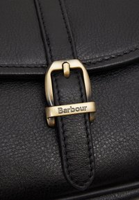 Czarna skórzana torba z teksturowaną powierzchnią, z klamrą na zatrzask w kolorze złotym i wytłoczonym logo „Barbour” na pasku.