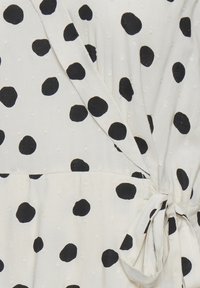 Tejido blanco con lunares negros, texturizado con patrones en relieve. Presenta un diseño envolvente con un detalle de lazo en la cintura.
