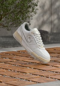 Balti odiniai sportbačiai su tekstūruotu paviršiumi, smėlio spalvos pado, apvaliu piršteliu ir ryškiu logotipu šone. Į komplektą įeina raišteliai ir paminkštintas apykaklė.