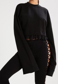 Felpa nera corta con maniche lunghe, caratterizzata da un orlo in pizzo arricciato. Abbinata a leggings neri con design a taglio incrociato.