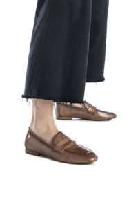 Mocasines de cuero marrón con un acabado brillante, que presentan un diseño de doble tira. Combinados con pantalones recortados de pierna ancha negros con dobladillos deshilachados.