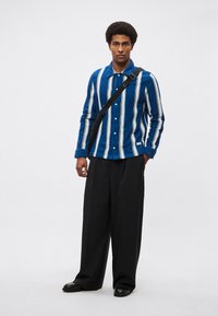 Niebiesko-biała koszula w paski na guziki z kołnierzykiem, połączona z luźnymi czarnymi spodniami i noszona z czarną torbą na ramię.