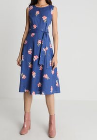 Blå blommig klänning med en figursydd överdel, ärmlös design och midjeband. Har rosa blommönster och en utsvängd kjol. Buren med rosa stövlar.
