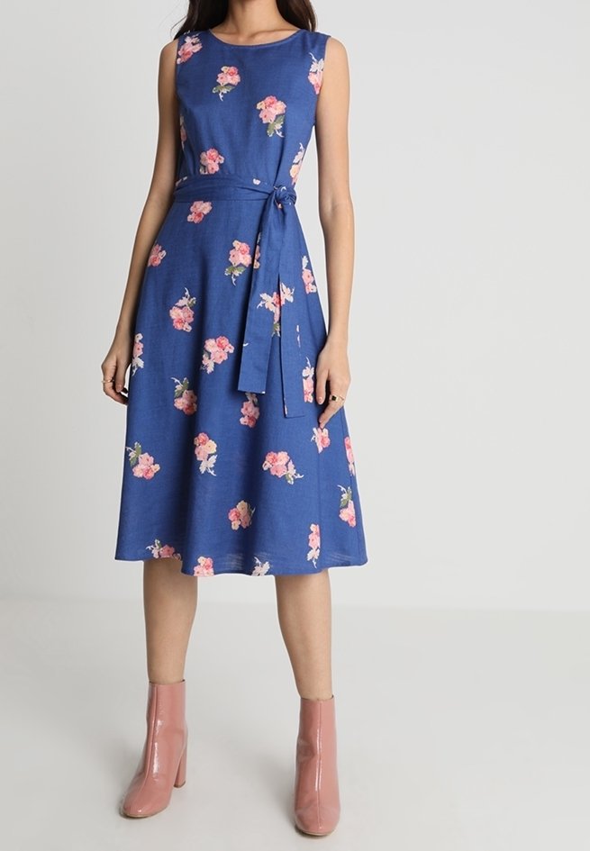 Blå blommig klänning med en figursydd överdel, ärmlös design och midjeband. Har rosa blommönster och en utsvängd kjol. Buren med rosa stövlar.