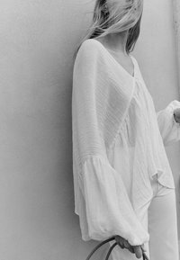 Blouse blanche texturée avec des manches évasées, dotée d'un col en V et d'une silhouette fluide, associée à un pantalon clair et un sac tissé.