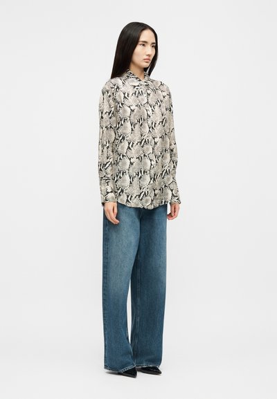 Woman standing wearing long-sleeve snake print blouse, wide-leg blue jeans, and black pointed toe shoes on a plain white background.