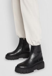 Zwarte leren enkellaarsjes met een dikke, geribbelde zool. Voorzien van een zijrits en een gladde textuur, gecombineerd met crèmekleurige broek.