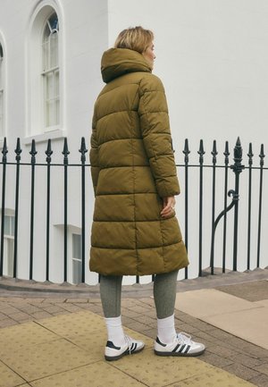Person steht auf dem Bürgersteig und trägt einen olivgrünen gepolsterten Mantel, graue Leggings, weiße Socken und weiße Sneaker mit schwarzen Streifen.