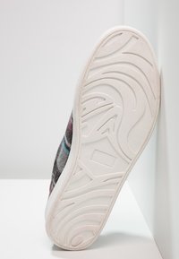 Semelle d'une chaussure à semelle en caoutchouc blanc avec des motifs de bande de roulement abstraits, positionnée verticalement contre un mur blanc et une surface blanche.