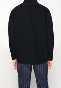 Chemise noire à manches longues avec col, poignets à boutons et ourlet droit. Portée avec un pantalon rayé marine. Le matériau semble lisse et mat.
