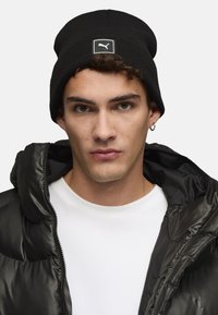 Gorro negro de canalé con un parche blanco del logo de Puma, combinado con una chaqueta negra tipo puffer y una camiseta blanca. Textura de tela suave.