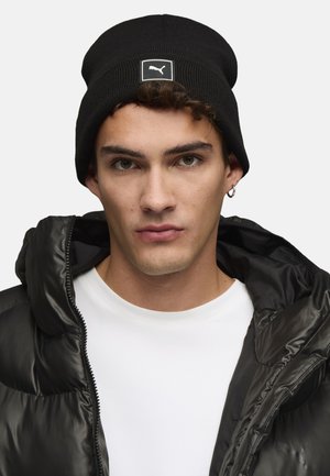Sort ribbet hue med et hvidt Puma-logo patch, båret sammen med en sort puffjakke og en hvid t-shirt. Glat stoftekstur.