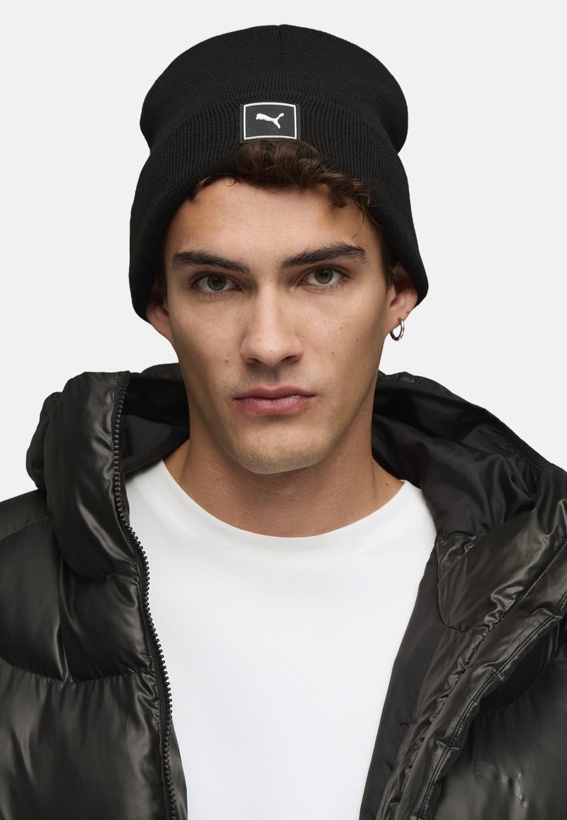 Gorro negro de canalé con un parche blanco del logo de Puma, combinado con una chaqueta negra tipo puffer y una camiseta blanca. Textura de tela suave.